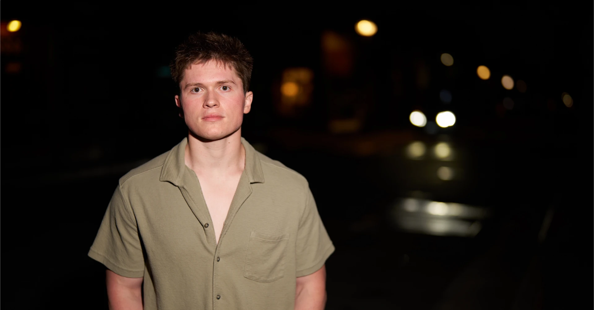 Jason Kushy night urban press photo in an olive button-down shirt with city lights bokeh