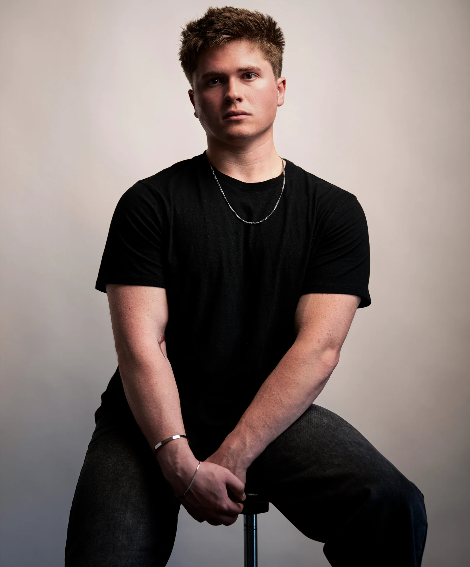 Seated studio portrait of Jason Kushy wearing a black t-shirt and chain necklace